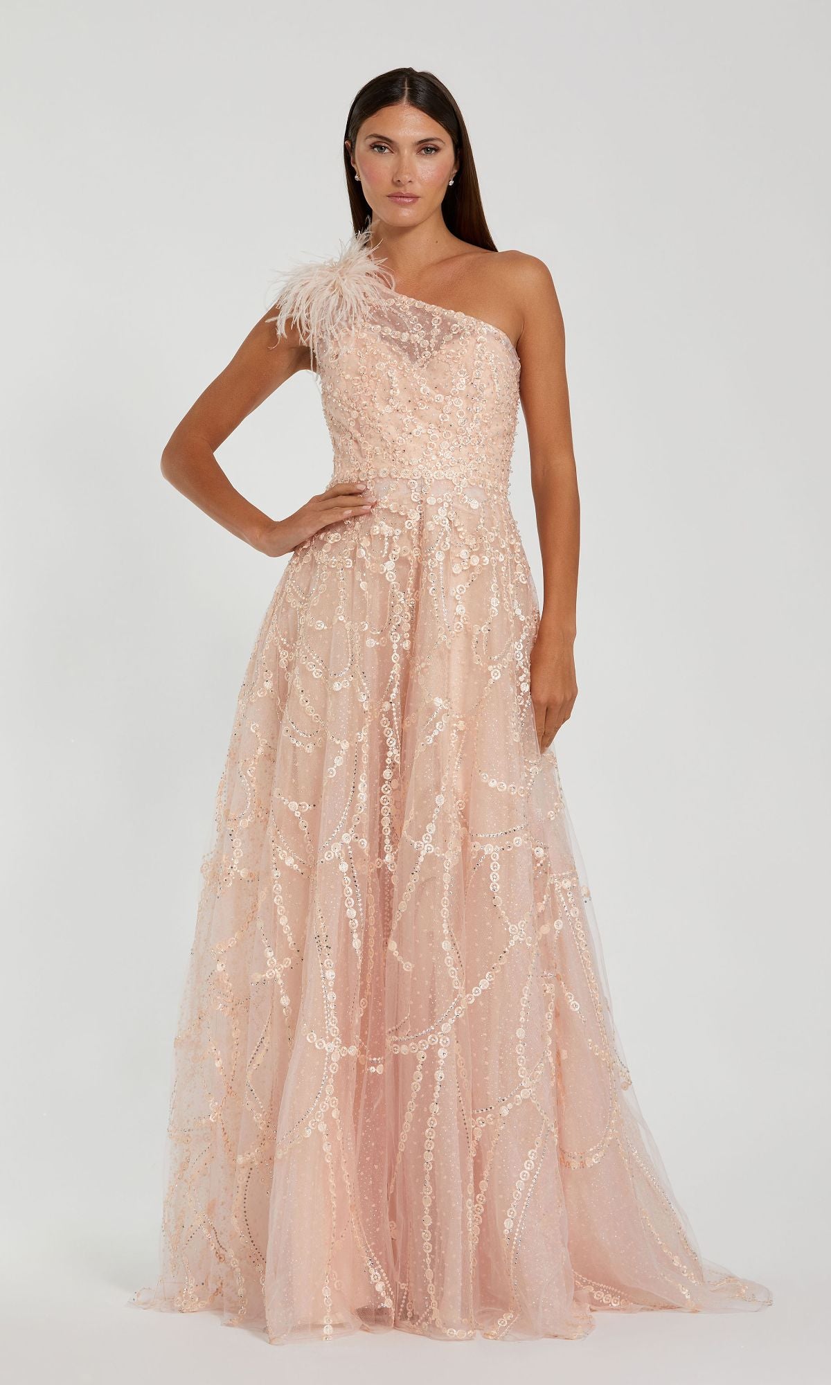 A woman stands elegantly against a plain light background in the Beaded One-Shoulder Prom Gown: Mac Duggal 20389, featuring blush pink fabric, beaded accents, and feather details.